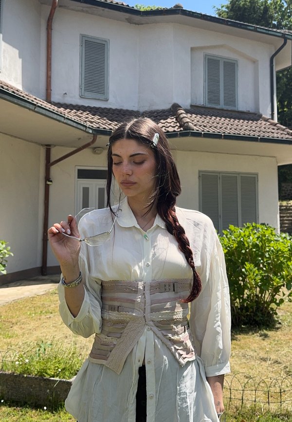 Young woman with long braided hair wearing a white blouse and beige corset, holding sunglasses, standing outside a house with closed shutters.