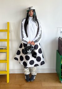 Žena oblečená v bílé bundě Adidas, černé a bílé sukni s puntíky, sladěných kalhotách, černých botách Adidas, proužkované čepici a držící černou tašku Adidas.