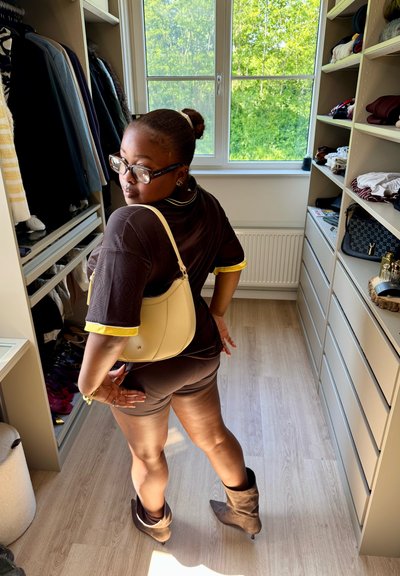 Mujer con gafas, atuendo negro, bolso beige y botas marrones posando en un vestidor luminoso con ropa y estantes.