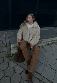 Ψεύτικη γούνα μπεζ σακάκι πάνω από ένα κρεμ πλεκτό πουλόβερ, συνδυασμένο με καφέ παντελόνι wide-leg και καφέ μπότες μέχρι τον αστράγαλο. Ομαλές, άνετες υφές είναι ορατές.