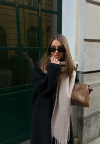 Femme en manteau noir, écharpe beige et lunettes de soleil, debout près d'une fenêtre verte et d'un mur en pierre beige, tenant un sac en cuir marron.