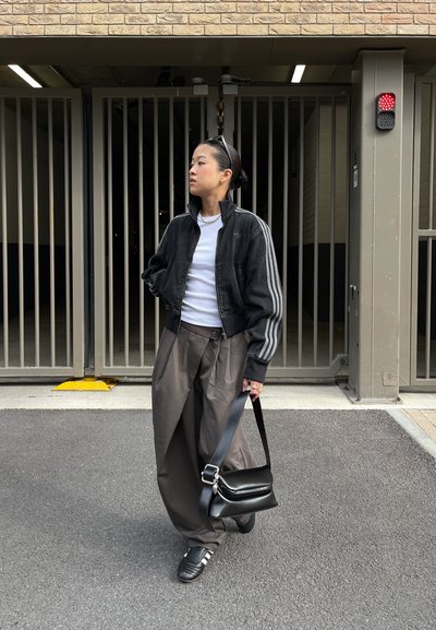 Chaqueta bomber negra con rayas grises, camisa blanca, pantalones anchos grises y bolso negro. Zapatillas negras con rayas blancas. Ambiente urbano.