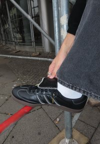 Svart skinnsneaker med hvite sømmer og gull detaljer, med snøring og en teksturert tunge, plassert mot en grå bakgrunn.