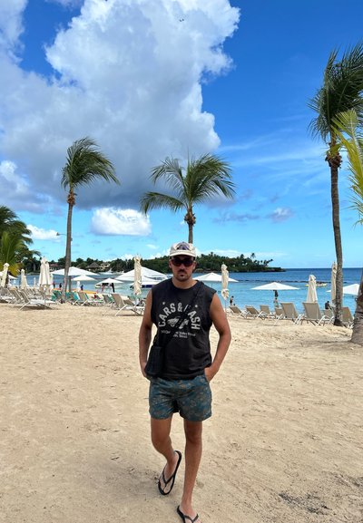 Camisa sin mangas negra con gráfico, pantalones cortos con un patrón en azul y marrón, chanclas y una gorra ligera, de pie en una playa de arena.