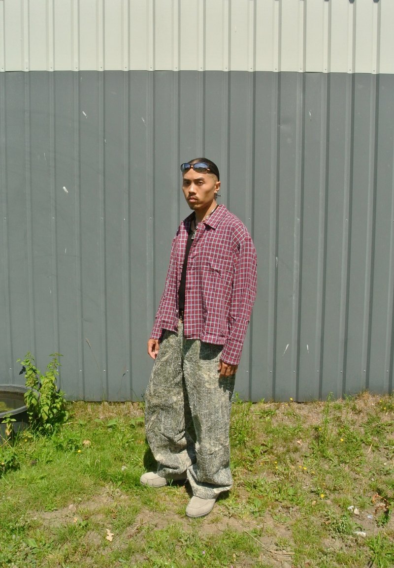 Homme portant des lunettes de soleil, une chemise à carreaux rouge et un pantalon gris ample, debout sur l'herbe devant un mur métallique gris.