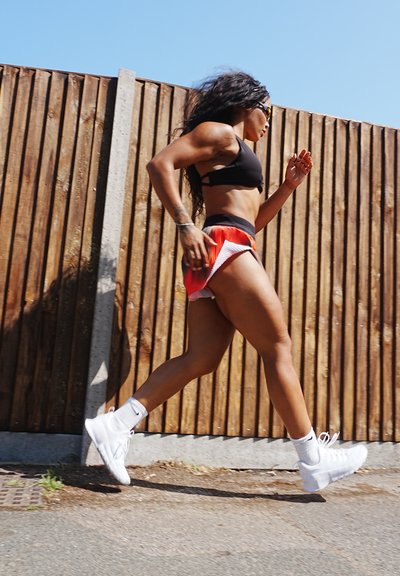 Sujetando un sujet dorado en una serie negra de entrenamiento, pantalones cortos rojos y blancos, junto con zapatillas blancas. La persona está corriendo junto a una cerca de madera con un cielo azul despejado arriba.