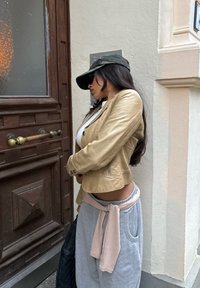 Light beige leather jacket, grey sweatpants with a pink tie waist, black hat, and sunglasses, standing beside a wooden door with a glass panel.