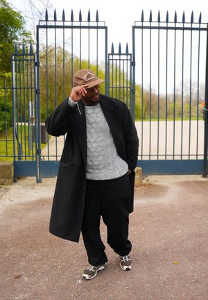 Una persona lleva un abrigo negro sobre un suéter gris, pantalones negros y una gorra marrón. Está de pie frente a una puerta azul.