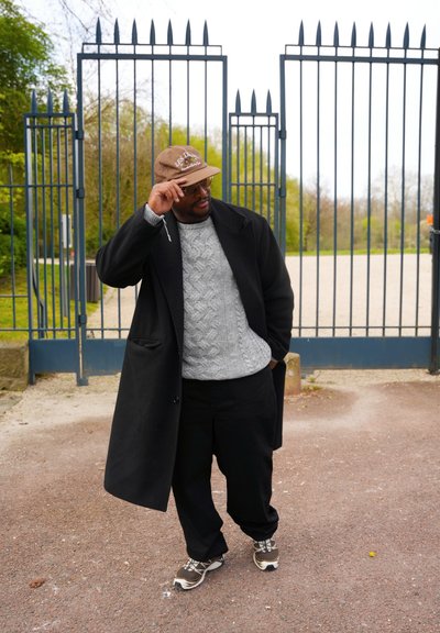 Una persona lleva un abrigo negro sobre un suéter gris, pantalones negros y una gorra marrón. Está de pie frente a una puerta azul.