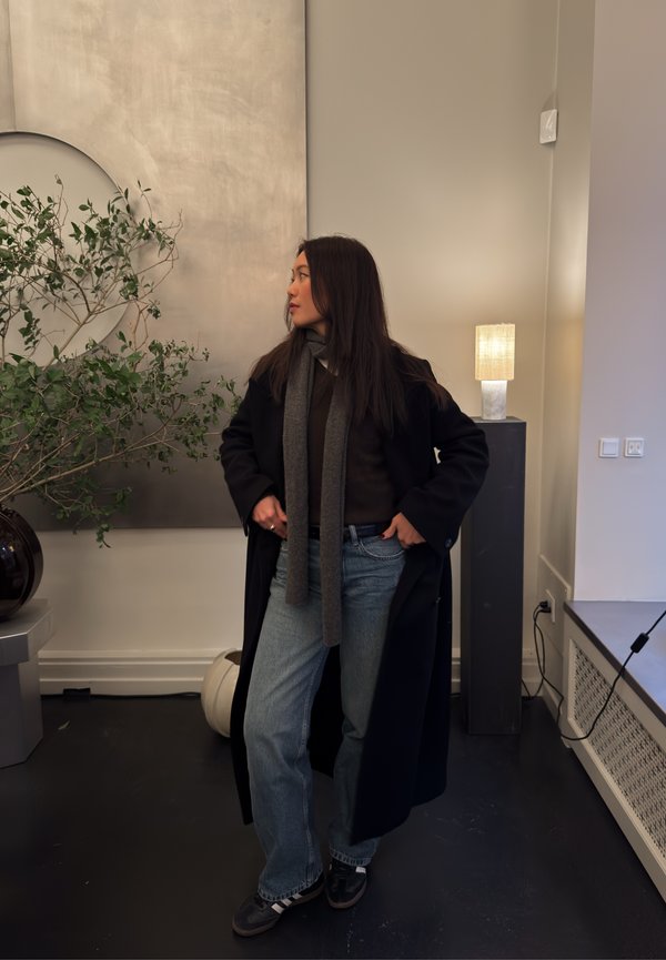 Frau in einem langen schwarzen Mantel, grauem Schal und Jeans steht drinnen in der Nähe einer grünen Pflanze und einer eingeschalteten Tischlampe auf einem Podest.