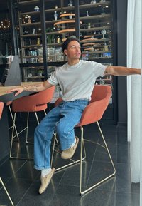Orange bezogenes Barhocker mit goldenen Beinen; Person in einem weißen T-Shirt und blauen Jeans, die an einem Holztisch in einem modernen Interieur sitzt.