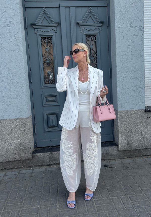 Femeie în sacou alb cu dungi fine și pantaloni transparenți brodați stă pe trotuarul gri în fața unei uși decorative albastre, ținând o geantă roz.