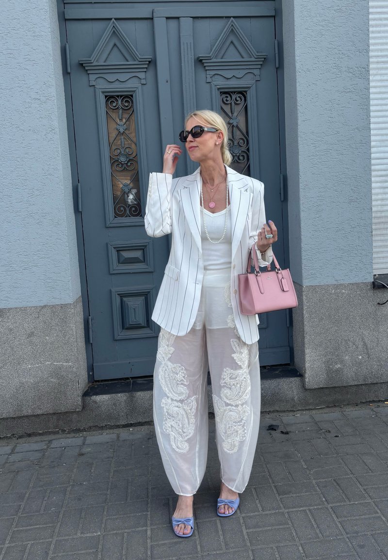 Femme en blazer blanc à fines rayures et pantalon transparent brodé, debout sur un trottoir gris devant une porte décorative bleue, tenant un sac à main rose.
