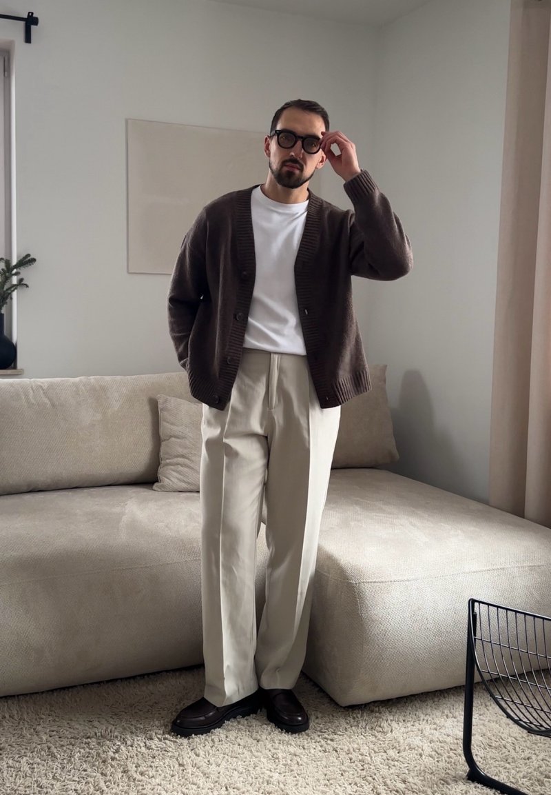 Uomo che si sistema gli occhiali tondi neri, indossa un cardigan marrone, camicia bianca, pantaloni beige, in piedi in un soggiorno moderno beige.