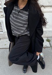 Um casaco preto sobre uma camisa polo riscada em preto e branco. Calças pretas, sapatos pretos brilhantes e uma bolsa trançada. Pavimento de cimento com vegetação.