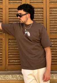 Braunes T-Shirt mit kurzen Ärmeln und einem Grafikdesign eines Flamingos auf der Brust, kombiniert mit geschichteten silbernen Halsketten und beigen Hosen. Texturierten Holz Hintergrund.