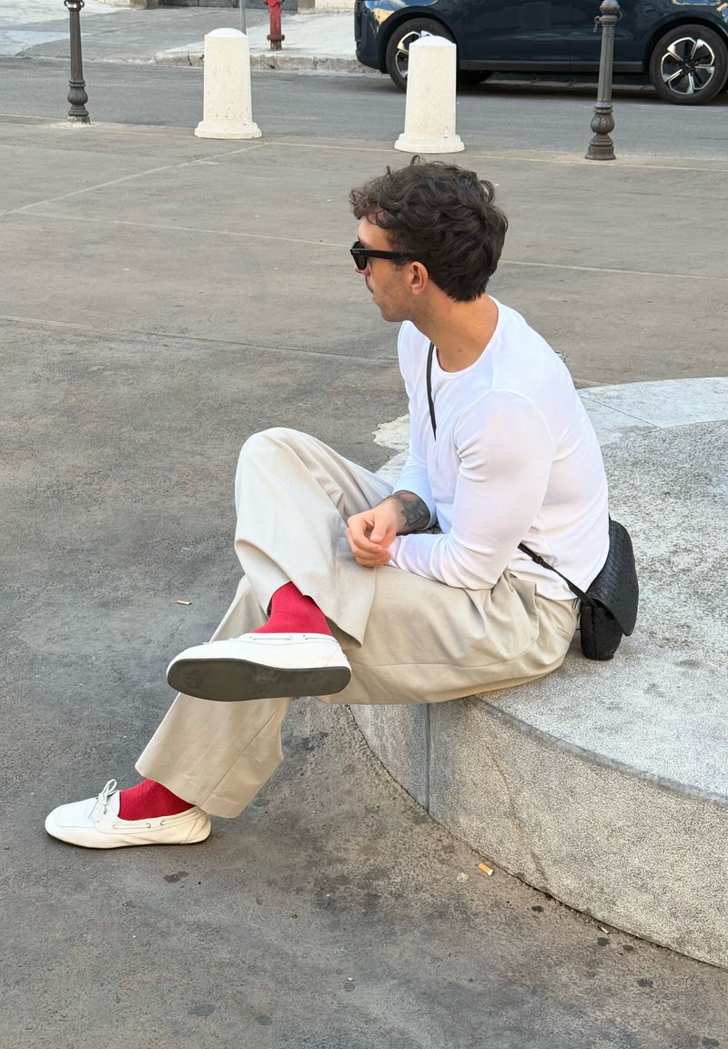 Mężczyzna w białej koszuli z długim rękawem, beżowych spodniach, czerwonych skarpetkach i białych butach siedzi na okrągłej kamiennej ławce na zewnątrz.