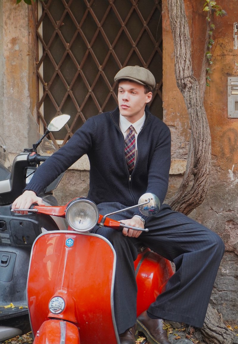 Rode vintage scooter met chroomaccenten en ronde koplamp; man in een donkere trui en geruite das zit casual, met een grijze pet op.