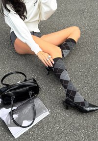 Fekete és szürke argyle mintás, térd fölé érő zoknik fekete magassarkú cipővel, fekete kézi táskával és magazinnal egy texturált felületen.