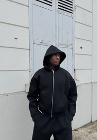 Juodas zip-up džemperis pagamintas iš storos medžiagos, su gobtuvu ir priekinėmis kišenėmis. Poruojamas su derančiomis sportinėmis kelnėmis, prieš neutralų foną.
