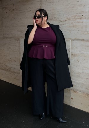 Modelo de talla grande con una blusa peplum en color ciruela, pantalones anchos negros y un abrigo largo negro, llevando gafas de sol negras y pendientes de aro dorados.