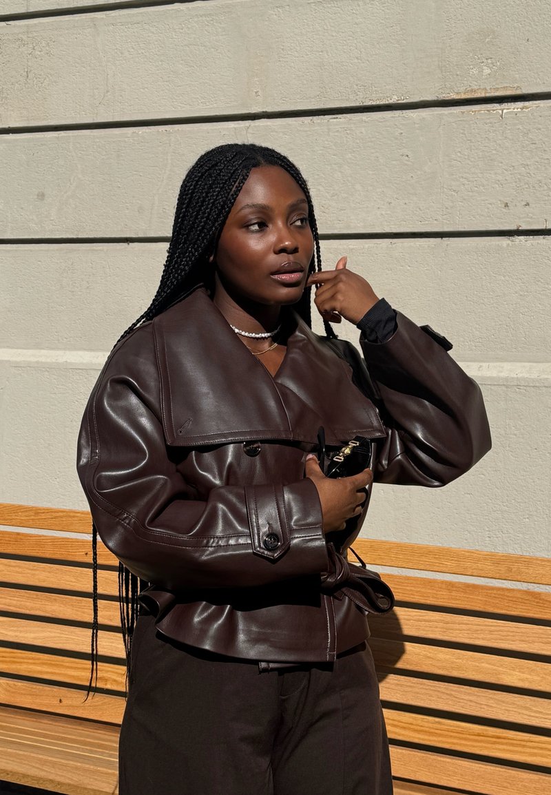 Femme aux longues tresses ajuste sa boucle d'oreille tout en portant une veste en cuir brun foncé et un collier de perles, debout près d'un banc en bois et d'un mur en béton.