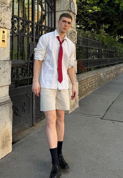 Camisa blanca de manga larga, corbata roja con un patrón, pantalones cortos beiges, calcetines negros y zapatos negros. De pie en una acera cerca de una entrada cerrada.