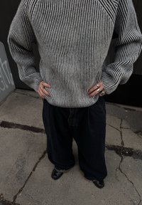 Grey and black striped knit jumper with long sleeves, paired with loose dark denim trousers and black shoes. Hands on hips, wearing rings.