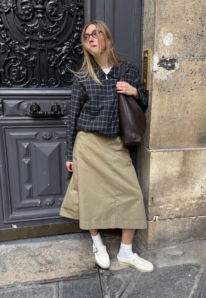 Femme aux longs cheveux blonds portant des lunettes, une veste à carreaux, une jupe midi beige, des chaussettes blanches et des chaussures, debout contre une porte décorative gris foncé.