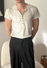 Krämfärgad kortärmad henley-tröja med ett marmorliknande mönster och fem knappar, ihop med svarta plisserade byxor.