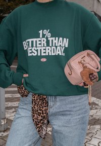 Zelená mikina s bílým nápisem "1% LEPŠÍ NEŽ VČERA", oprané světle modré džíny a malá růžová kabelka s hnědými detaily.