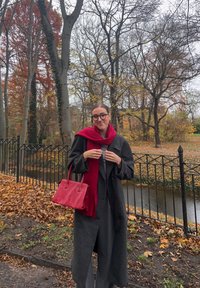 Siv plašč, rdeč šal in rdeča torbica. Drevesa z jesenskimi listi in vidna vodna površina v ozadju.