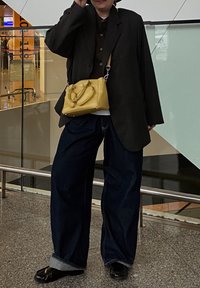 Svart oversized blazer, brun cardigan, hvit skjorte, vide mørk denimjeans, svarte sko, og en gul håndveske med dobbelthåndtak.