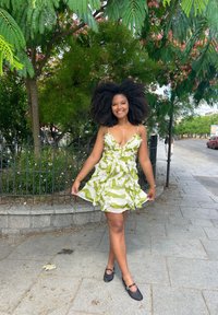 Vestido de rayas verdes y blancas con volantes, escote en V y tirantes finos, combinado con zapatos planos negros en una acera rodeada de vegetación.