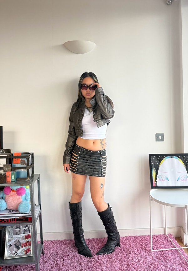 Femme portant une veste en cuir courte, un haut blanc, une mini-jupe noire à découpes, de hautes bottes noires et des lunettes de soleil, debout sur un tapis rose à l'intérieur.