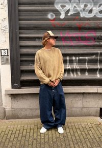 Ryškiai didelio dydžio smėlio spalvos megztinis su plačiomis rankovėmis, derinamas su laisvomis tamsiais džinsais ir baltais sportbačiais. Stovi prie tekstūruotos sienos.