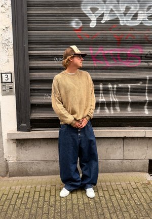 Suéter beige de punto grueso y oversize con mangas anchas, combinado con jeans de mezclilla oscuros y holgados y zapatillas blancas. De pie contra una pared texturizada.