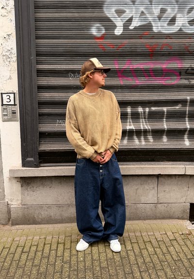 Suéter beige de punto grueso y oversize con mangas anchas, combinado con jeans de mezclilla oscuros y holgados y zapatillas blancas. De pie contra una pared texturizada.