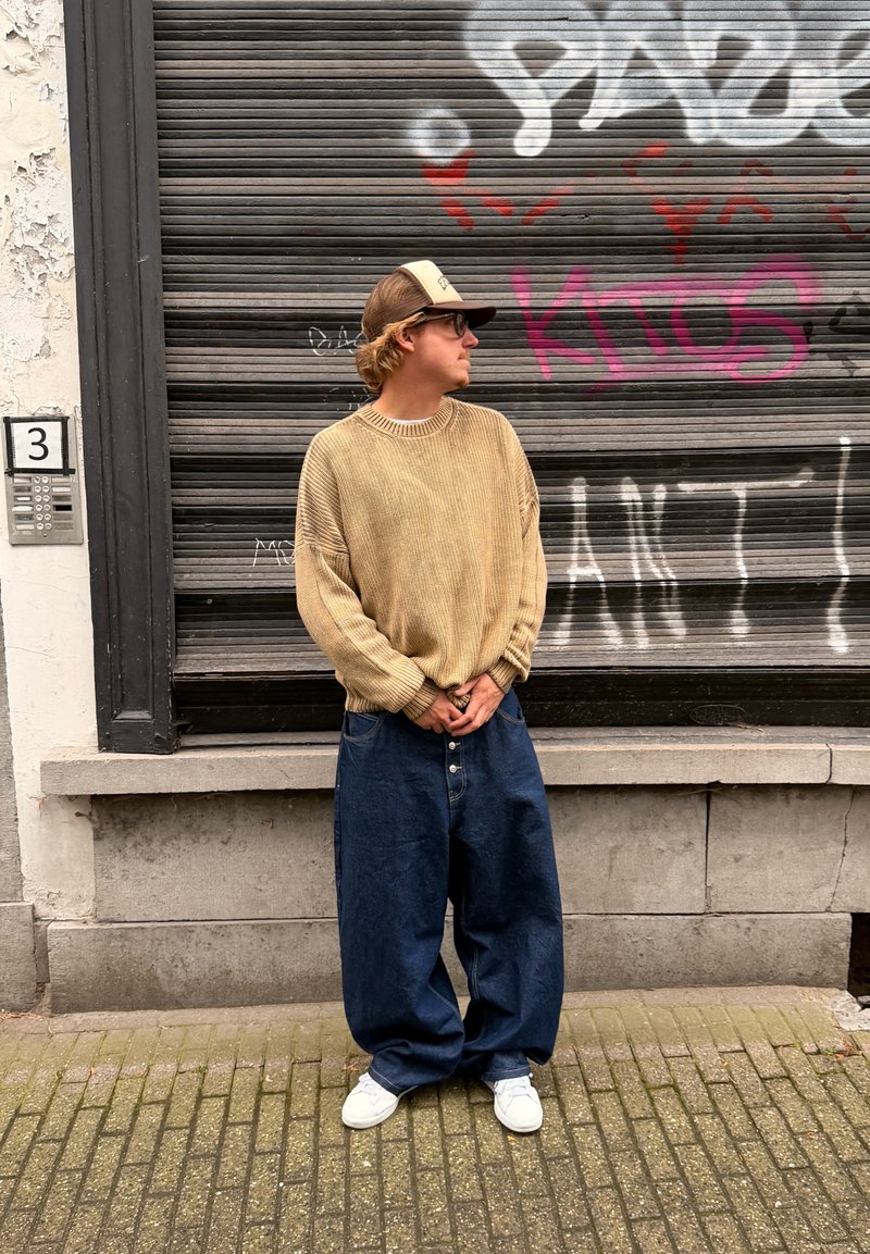 Maglione beige oversize a coste con ampie maniche, abbinato a jeans in denim scuro larghi e sneaker bianche. In piedi contro una parete texturizzata.