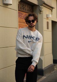 Φανέλα Nike σε off-white απόχρωση με oversized εφαρμογή, διακοσμημένη με μπλε λογότυπο. Μαύρο παντελόνι, γυαλιά ηλίου και ένα ασημένιο δαχτυλίδι προσθέτουν λεπτομέρειες στην εμφάνιση.