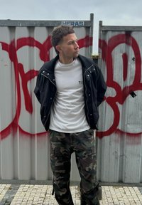 Giacca nera con zip, t-shirt bianca e pantaloni cargo in camouflage. Sullo sfondo, recinzioni metalliche grigie con accenti di graffiti rossi.