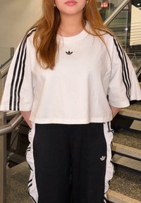 T-shirt blanc coupé court avec logo adidas noir, associée à un pantalon noir avec des bandes blanches sur les côtés et des accents froncés.