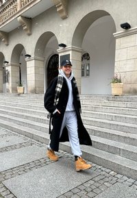 Čierny kabát, šedá mikina a tepláky. Hnedé čižmy so šnúrkami. Škvrnitý šál. Kamenné schody a architektúra v pozadí, padajúce ľahké sneženie.