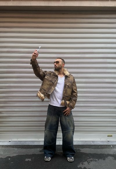 Chaqueta de cuero marrón corta con detalle de piel, camisa blanca y jeans oscuros de pierna ancha. La persona sostiene un teléfono mientras posa.