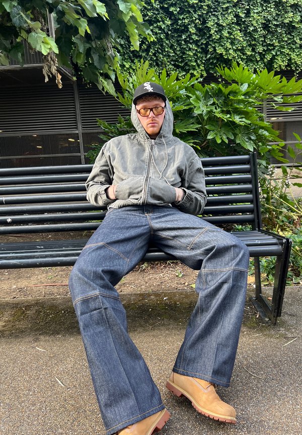 Graue Reißverschlussjacke mit großen Taschen, blaue Jeans mit sichtbaren Nähten und braune Arbeitsstiefel auf einer Parkbank mit grüner Vegetation im Hintergrund.