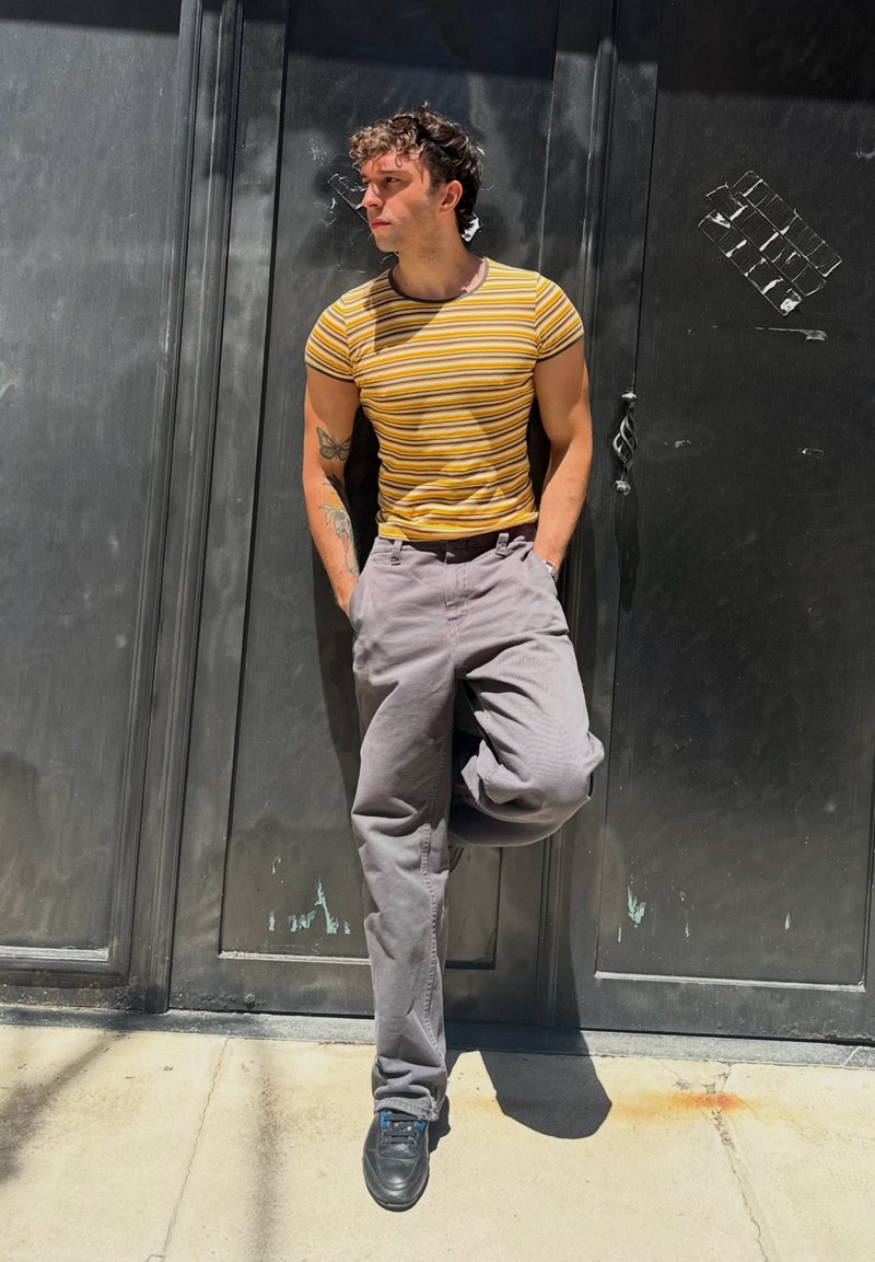 Jeune homme aux cheveux bouclés portant une chemise jaune rayée et un pantalon gris, appuyé contre une porte sombre, les mains dans les poches, regardant vers sa gauche sous le soleil.