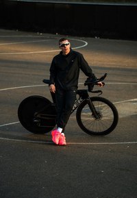 Homme en tenue de sport noire et chaussures rose vif appuyé sur un vélo de course noir sur un terrain extérieur sous une lumière solaire basse.