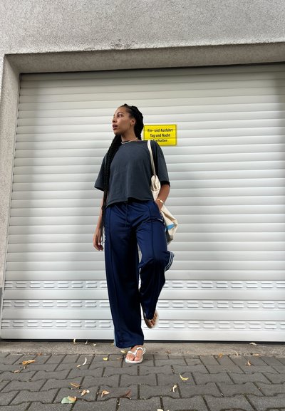 Camiseta corta gris, pantalones anchos azul marino con rayas laterales y chanclas blancas. Bolsa de mano beige con detalles azules. Fondo gris.