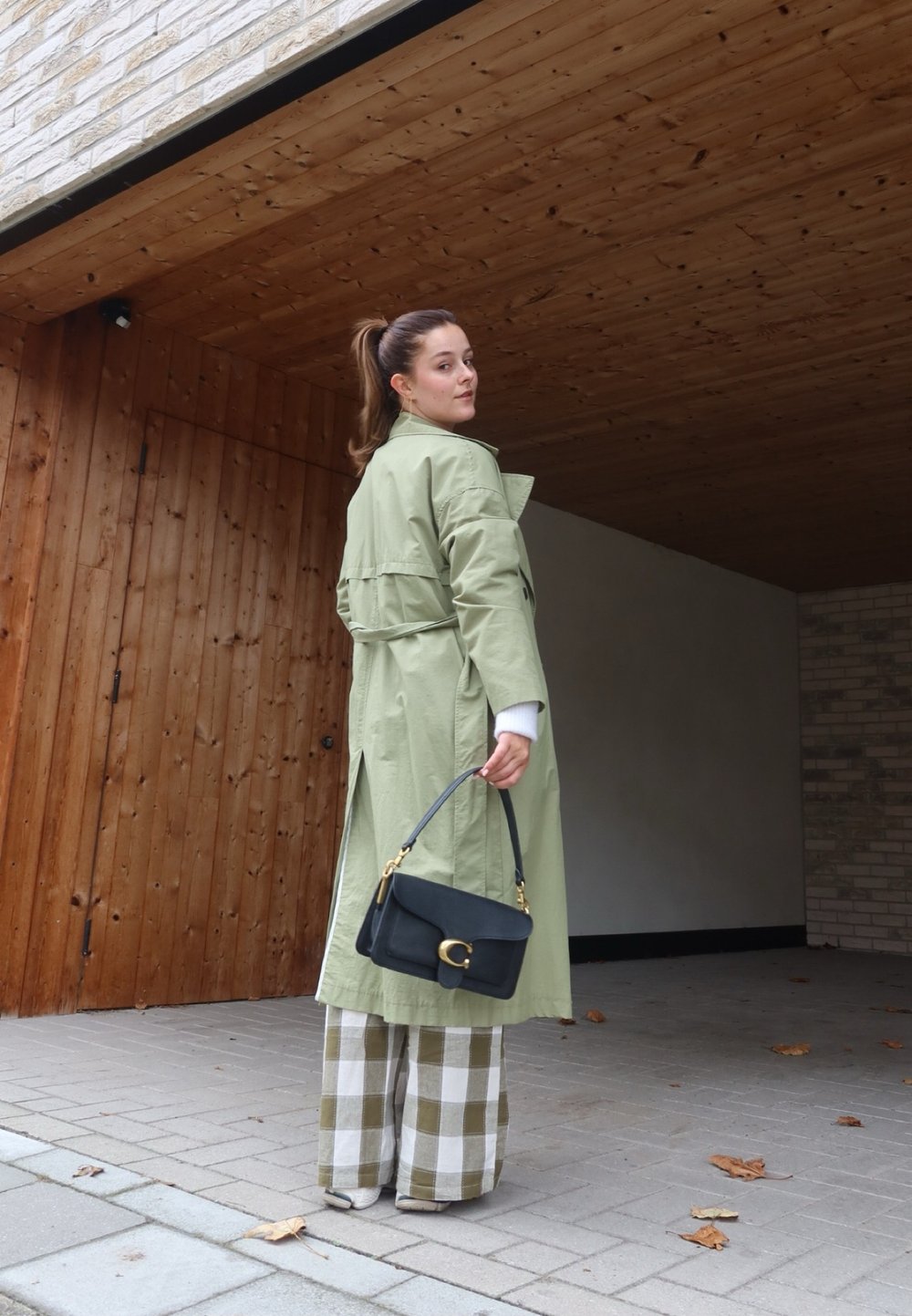 Ανοιχτό πράσινο παλτό με ζώνη, μαύρη τσάντα χειρός με χρυσά αξεσουάρ και καρό παντελόνια σε πράσινες και λευκές αποχρώσεις.