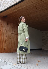 Ανοιχτό πράσινο παλτό με ζώνη, μαύρη τσάντα χειρός με χρυσά αξεσουάρ και καρό παντελόνια σε πράσινες και λευκές αποχρώσεις.